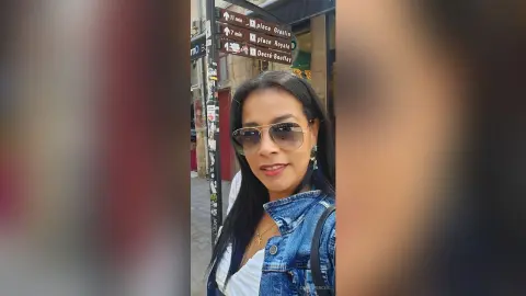 Smiling woman in sunglasses and denim jacket walking on a sunny city street near historic street signs.