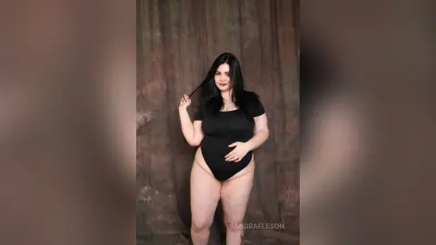 Smiling woman in a black bodysuit posing against a textured brown backdrop in a studio setting.