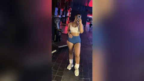 Athletic woman in white sports bra and blue shorts taking a mirror selfie in a moody, dimly lit gym.
