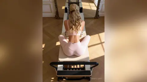 Blonde woman in pink yoga wear performing seated stretches on a Pilates reformer in a sunlit studio.