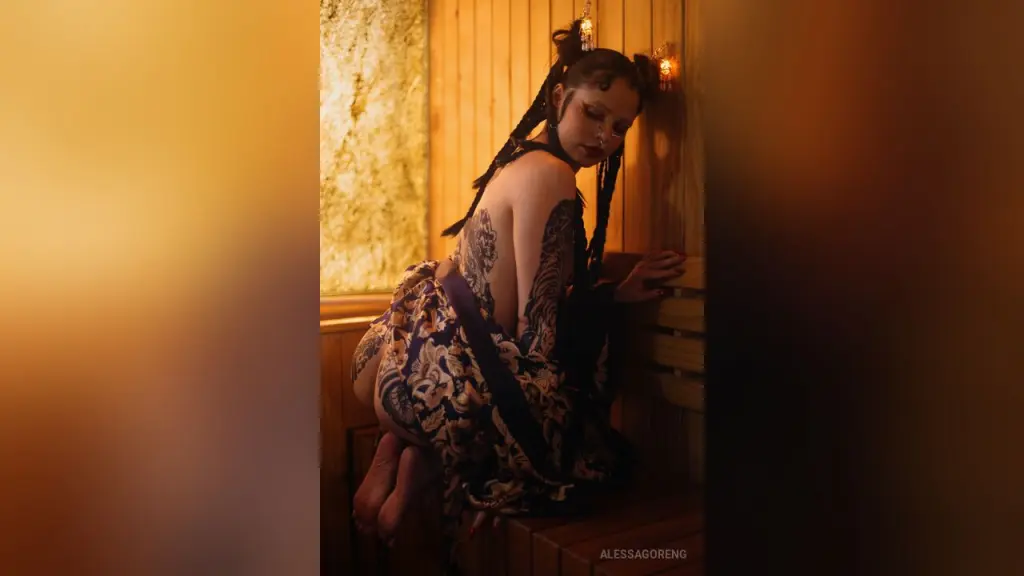 AlessaGoreng — Tattooed woman in a floral wrap sitting in a warm, moody wooden sauna.