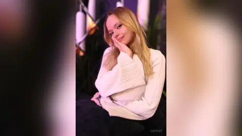 LucyLoy smiles at the camera while wearing a white long-sleeve shirt and resting her chin on her hand.