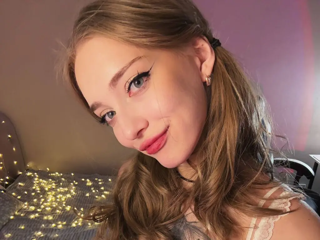 Smiling young woman with blonde hair and winged eyeliner posing in a cozy bedroom with fairy lights.