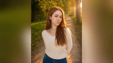 Young woman in a white knit sweater standing on a sunlit forest path during golden hour.