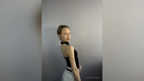 MalliaLucioni poses sideways against a grey wall, wearing a black halter top and white shorts, looking back over her shoulder.