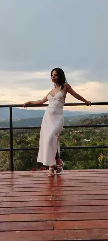 Elegant woman in a white satin slip dress posing on a wooden balcony overlooking a lush green mountain landscape.