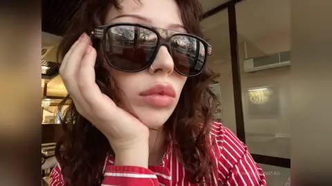 Young woman with wavy hair wearing dark sunglasses and a red striped shirt in a bright indoor setting.