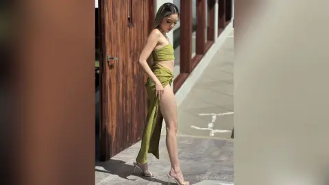 Elegant woman in a green slit maxi dress posing outdoors by a rustic wooden door in bright sunlight.