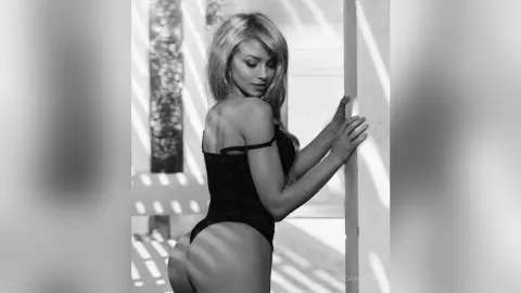 Blonde woman in black lace bodysuit posing by a sunlit window in a moody black and white portrait.