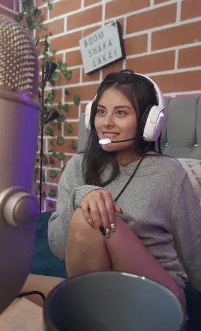 Smiling young woman with headphones and microphone recording a podcast in a cozy room with brick walls.