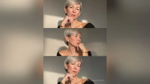 BellaMontclaire poses with short silver hair and a black top, looking thoughtfully into the distance against a soft-lit wall.