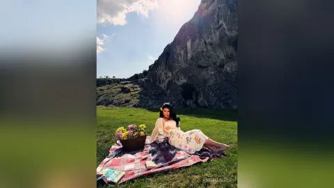 Woman in floral dress having a picnic on a blanket near a large mountain under a sunny sky.