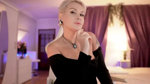 Elegant woman in a black off-the-shoulder dress posing in a cozy, dimly lit bedroom with purple curtains.