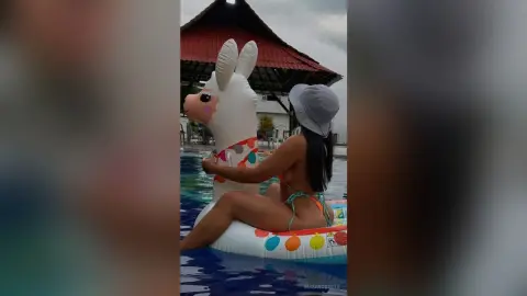 Woman in a bikini relaxing on a large white llama floatie in a swimming pool on a bright summer day.