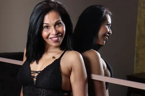 Smiling woman in a black lace outfit looking into a mirror in a moody indoor setting.