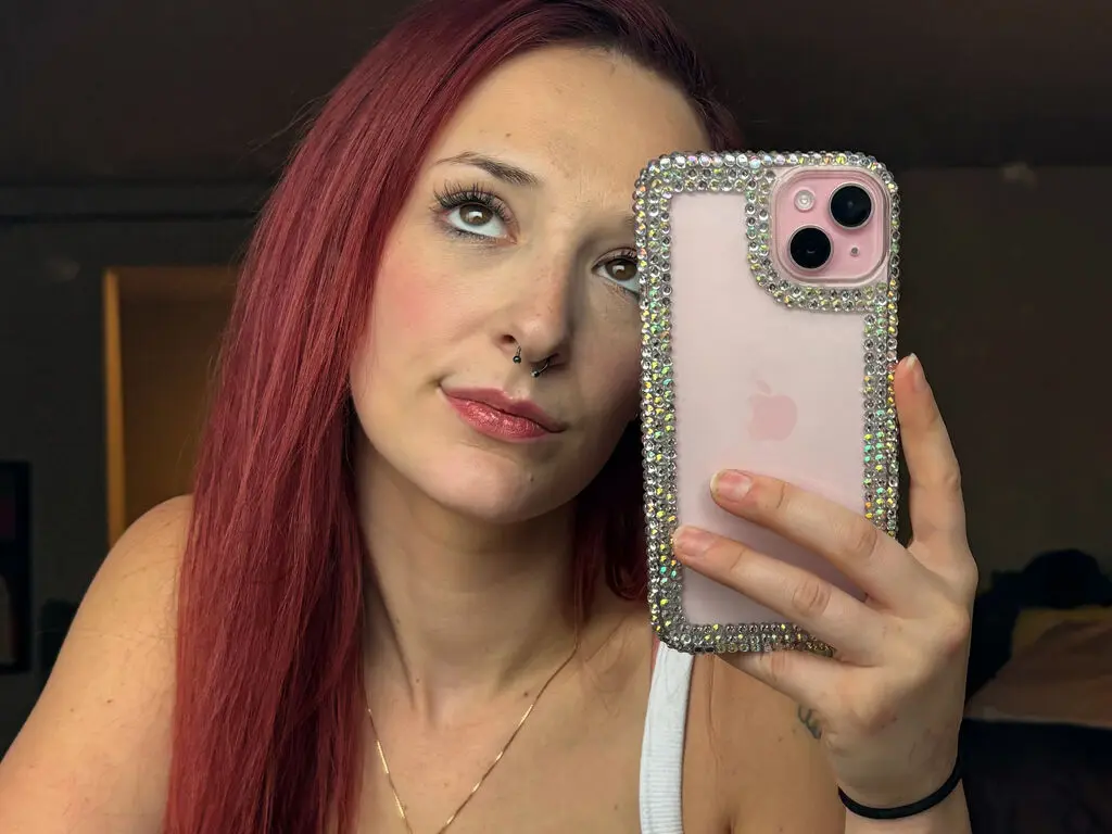 Red-haired woman with nose ring taking a selfie in a white tank top using a sparkly rhinestone phone case.