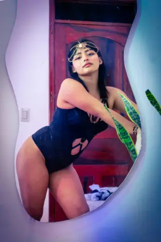 Young woman in a black bodysuit and gold headpiece posing confidently in a warm indoor setting.