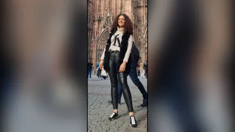 Smiling woman in a white sweater and black trousers standing before a historic gothic cathedral.