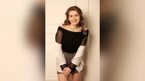Smiling young woman in a black sheer top and plaid skirt standing against a white door in a bright indoor setting.