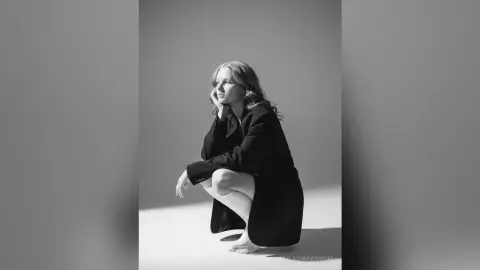 Melancholic woman in a black blazer sitting on a white floor in a minimalist studio setting.