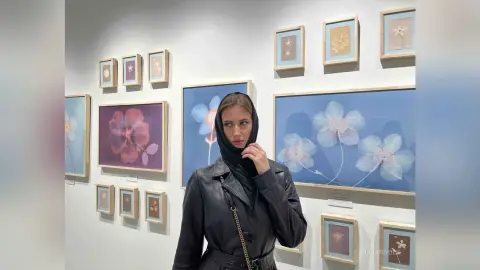 Woman in a black leather jacket and hijab standing in an art gallery surrounded by floral paintings.