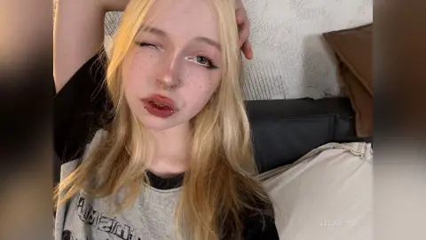Blonde woman with freckles and nose ring wearing a grey graphic t-shirt in a cozy indoor setting.