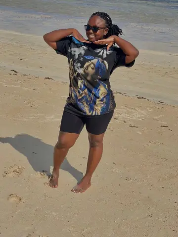 PrettyDoll22 stands on a sandy beach wearing a black graphic t-shirt, black shorts, and sunglasses, posing with her hands near her face.