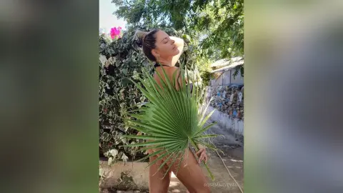 Young woman in black bikini holding a large green palm leaf in a sunny outdoor garden setting.