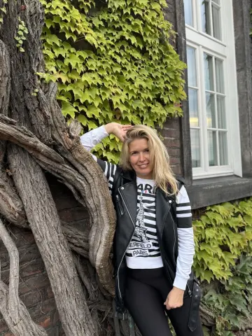 NikaZen is a blonde woman wearing a black and white striped shirt and a black vest, posing outdoors next to a large tree trunk and green ivy.