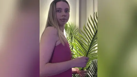 Blonde woman in a magenta tank top posing next to a green tropical plant in a bright indoor setting.