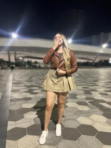 miarossexxXX wears a brown leather jacket and a tan pleated mini skirt with white sneakers while standing on a tiled outdoor walkway at night.
