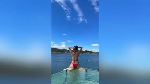 Woman in a red bikini relaxing on a poolside deck overlooking a calm blue ocean under a sunny sky.
