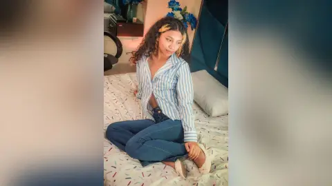 Young woman in a blue striped shirt and jeans sitting on a bed in a cozy, sunlit bedroom.