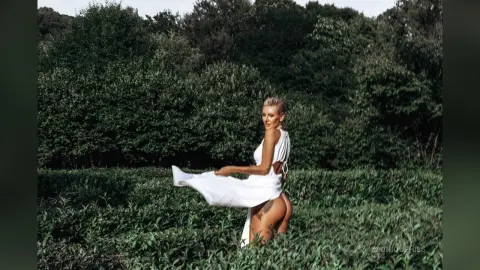 Blonde woman in a flowing white summer dress dancing in a lush green field under soft sunlight.