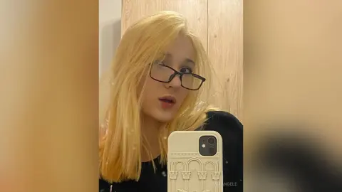 Blonde woman with glasses taking a selfie in a cozy indoor setting.