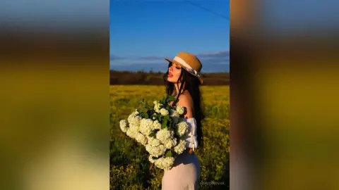 Dreamy woman in a white summer outfit and straw hat holding a large white hydrangea bouquet in a golden field.