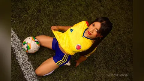 NairaWestbrook wears a yellow soccer jersey and blue shorts while sitting on a green grass field next to a soccer ball.