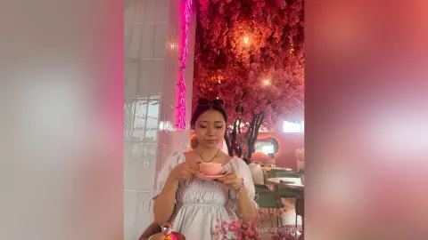 Young woman in a white summer dress enjoying tea in a dreamy pink floral cafe setting.