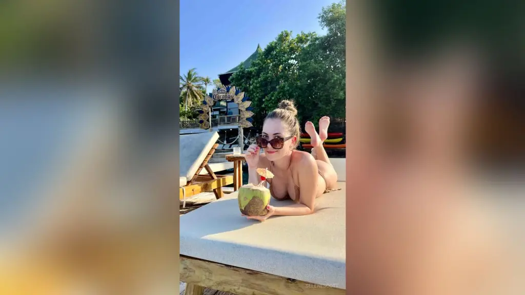 SeleneScott — Smiling woman in sunglasses relaxing on a sunbed with a coconut drink at a tropical beach resort.