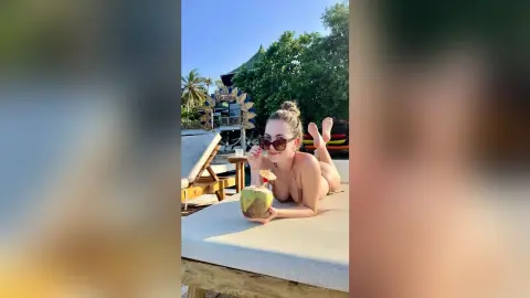 Smiling woman in sunglasses relaxing on a sunbed with a coconut drink at a tropical beach resort.