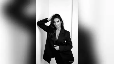 AnnaKarev poses in a black blazer and pants, leaning against a white wall with her hand in her long dark hair.