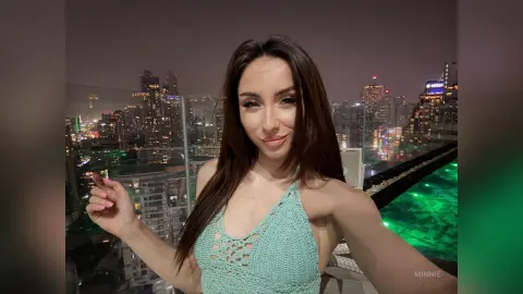Smiling woman in a mint green crochet halter top enjoying a night view from a rooftop pool overlooking a city skyline.