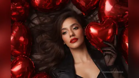 Glamorous woman in black outfit lying amidst shiny red heart balloons for a romantic Valentine's Day mood.