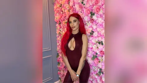 Red-haired woman in a sleek burgundy bodysuit posing against a romantic pink floral wall background.