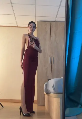 Elegant woman in a long burgundy evening gown posing in a bedroom mirror for a sophisticated look.