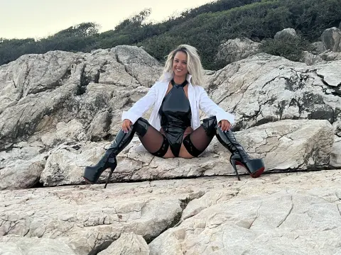 Smiling blonde woman in a black latex bodysuit and high heel boots sitting on coastal rocks at sunset.
