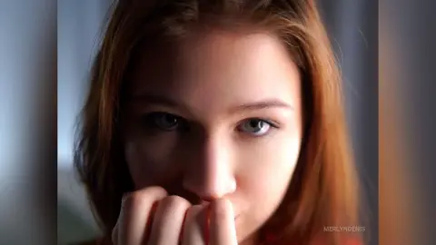 Close-up portrait of a young woman with red hair looking thoughtfully into the camera in soft lighting.