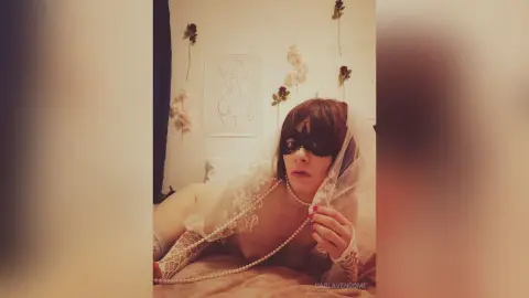 Woman in white lace lingerie and black masquerade mask posing on a bed in a dreamy, romantic setting.