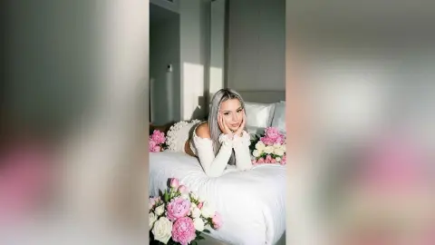 Kalira poses on a white bed with long silver hair, wearing a white lace top and resting her chin on her hands.