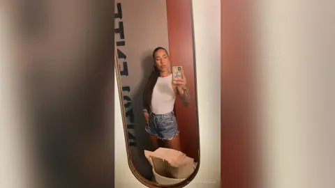 VictoriaFontaine poses for a mirror selfie wearing a white tank top and denim shorts, showing off her long dark hair and tattoos.